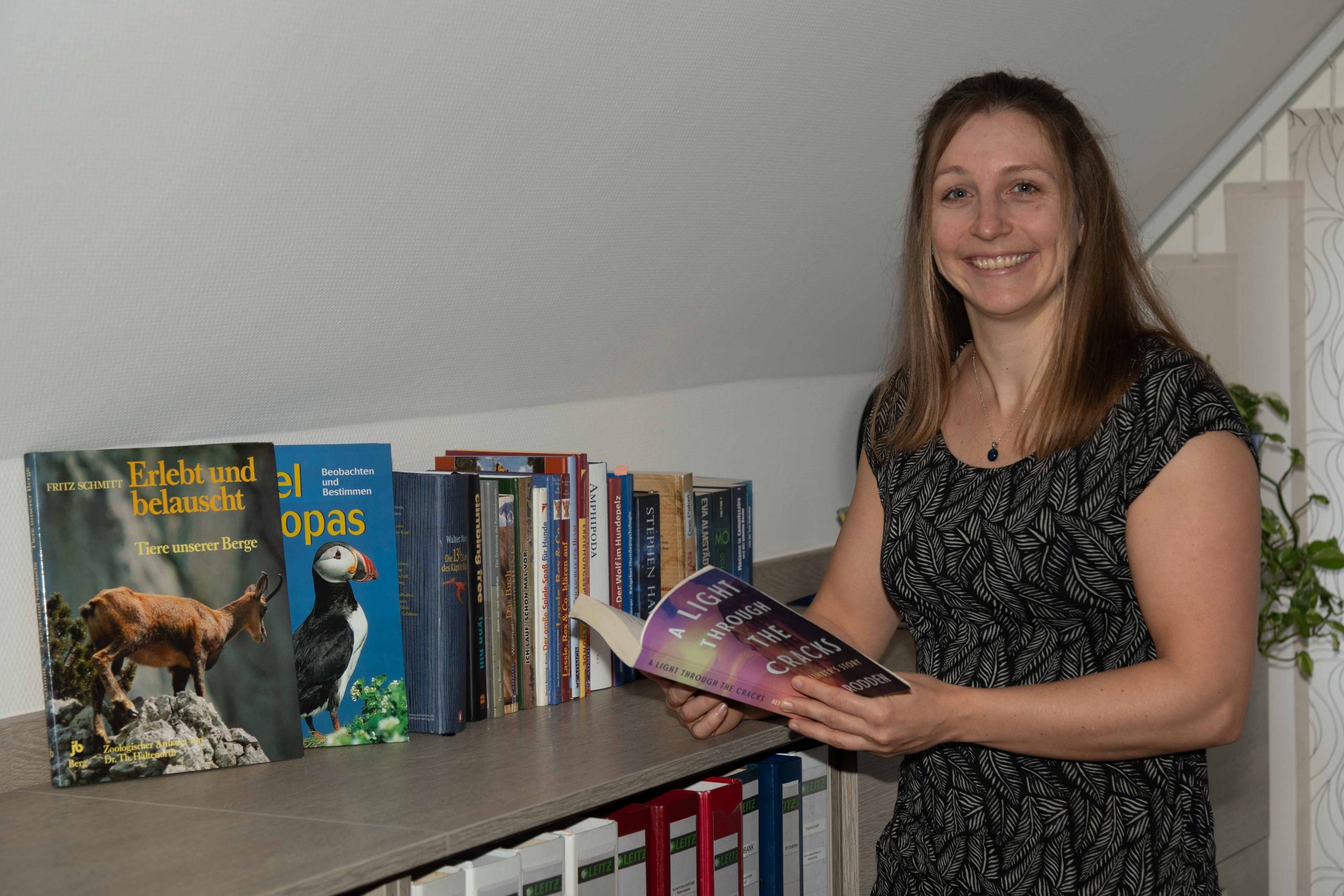 Dr. Martina Weiss mit dem Buch "A light through the Cracks", der Geschichte der bekannten Kletterin Beth Rodden, in der Hand vor einem Bücherregal mit Sachbüchern.