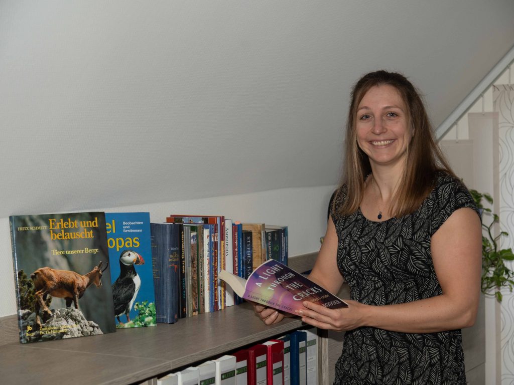 Dr. Martina Weiss mit dem Buch "A light through the Cracks", der Geschichte der bekannten Kletterin Beth Rodden, in der Hand vor einem Bücherregal mit Sachbüchern.