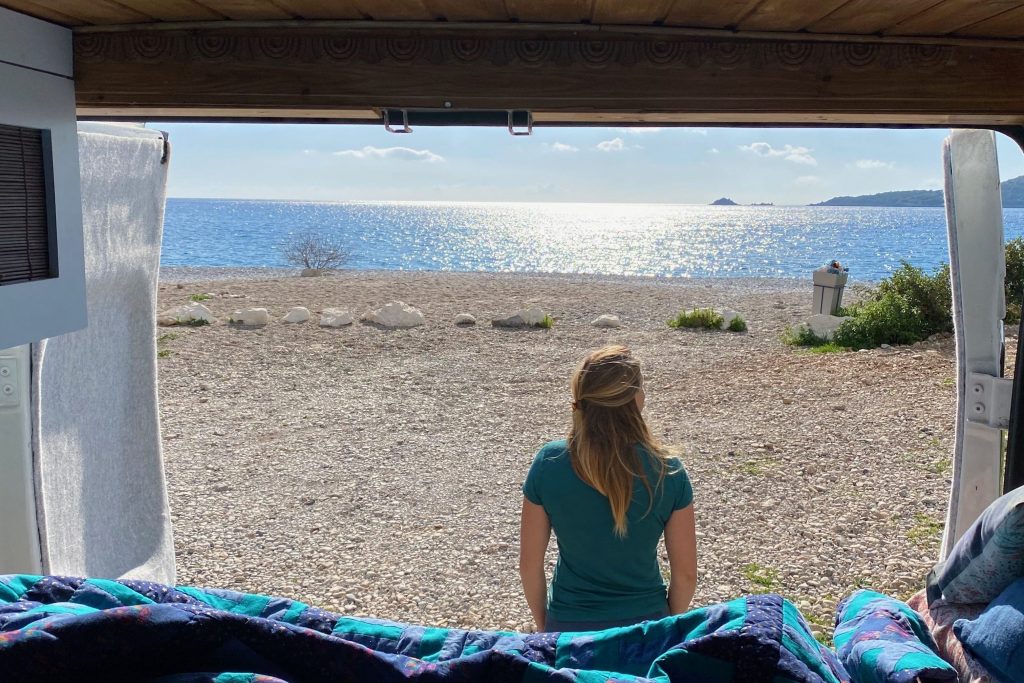 Blick aus dem zum Camper ausgebauten Lieferwagens an einem Strand in Kyparissi, Griechenland. Dr. Martina Weiss steht mit dem Rücken zur Kamera und schaut aufs Meer.