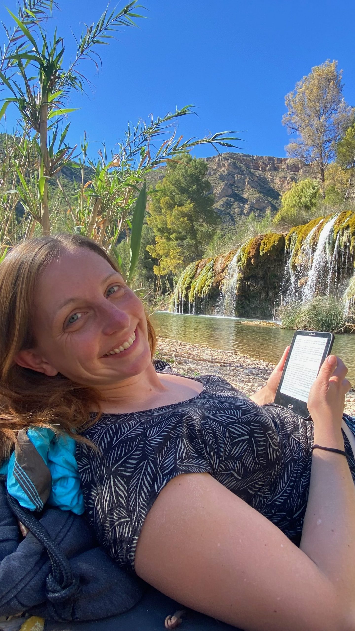 Dr. Martina Weiss beim Lesen eines eBooks an einem See mit Wasserfall in Spanien. 